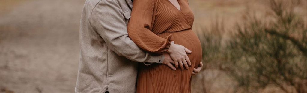 Vrouw is zwanger. Man omarmt de buik en staat achter haar.